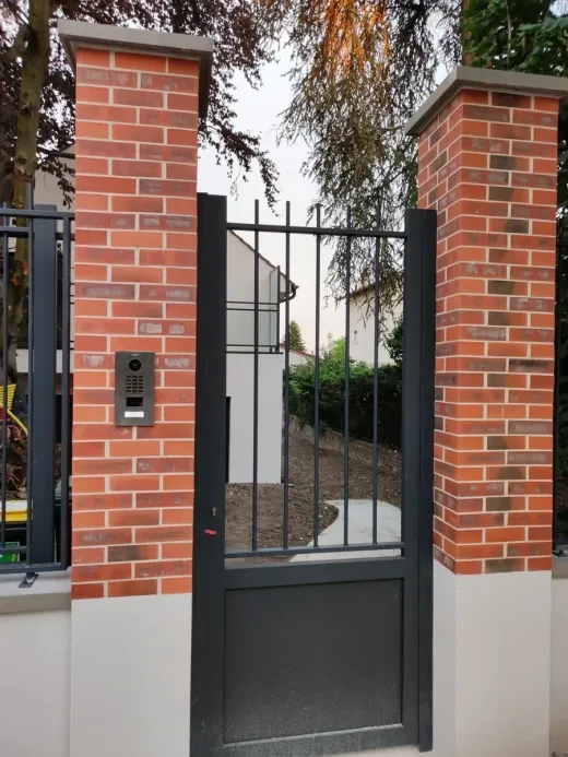 Platine de rue posée sur un pilier de brique, à côté d’un portail en fer